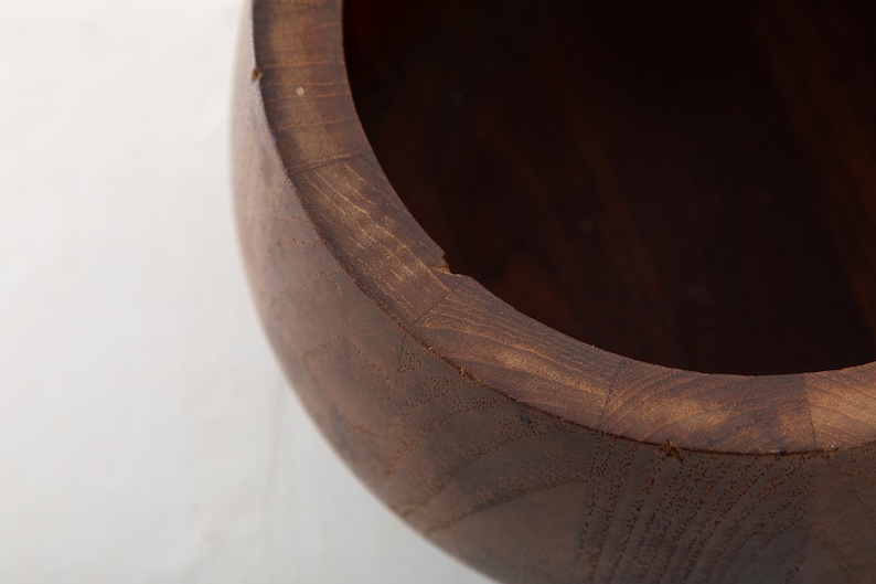 May include: Close-up of a wooden bowl with a dark brown finish. The bowl's rim is visible, showing the natural wood grain and a slight imperfection. The interior is dark, suggesting a deep cavity. This handcrafted bowl is suitable for kitchen use.