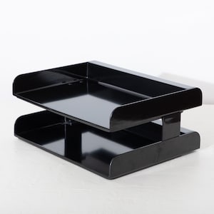May include: A glossy black two-tier desk organizer. The organizer has a rectangular shape with rounded edges and a connecting support in the center. The top and bottom trays are identical. The office supply item is made of a shiny material.