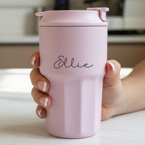 May include: A light pink reusable travel mug with a matte finish and a matching lid. The mug features a vertical ribbed texture on the lower half and the name Ellie printed in a black cursive script font on the centre of the smooth upper section.