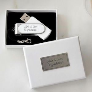 May include: A white USB flash drive with a silver metal accent featuring the text Marie & Jason Congratulations! inside a white gift box. The box lid has a rectangular silver metal plaque with the same text Marie & Jason Congratulations!