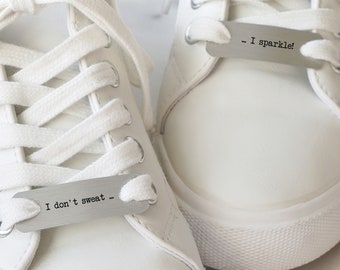 May include: A pair of white trainers featuring silver-toned metal shoelace tags. The left tag is engraved with the text 'I don't sweat ...' and the right tag is engraved with the text '... I sparkle!' threaded through the white shoelaces.