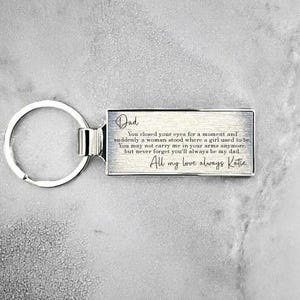 May include: Silver keyring with a rectangular tag engraved with a message to a dad from his daughter. The message reads: "Dad You closed your eyes for a moment and suddenly a woman stood where a girl used to be. You may not carry me in your arms anymore, but never forget you'll always be my dad. All my love always Katie"