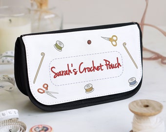 Puede incluir: Un estuche con cremallera en blanco y negro que presenta el texto Sarah's Crochet Pouch en letra roja. El panel frontal blanco está decorado con pequeñas ilustraciones de artículos de costura, como tijeras, carretes de hilo, botones y ganchillos.