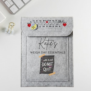 May include: A grey felt pouch with a flap, featuring the text "Katie's WEIGH DAY ESSENTIALS." The pouch has a progress tracker with hearts and percentages, and a note that says "DO NOT QUIT." A keyboard and binder clip are in the background.