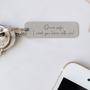 May include: Silver keyring with the text "Drive safe, I need you here with me!" on a white background.