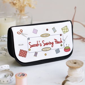 May include: A black and white sewing pouch with the text "Sarah's Sewing Pouch" in red. The pouch is decorated with cartoon images of sewing tools and fabric swatches. A measuring tape, thread spools, and scissors are in the foreground.