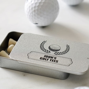 May include: A silver metal tin with a lid, containing golf tees. The lid has a black and white graphic of a golf ball with a laurel wreath and the text "John's Golf Tees".