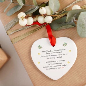 May include: A white heart-shaped ceramic Christmas ornament with a red ribbon, inscribed with a poem. The ornament is decorated with green sprigs and gold stars. It is placed on a wrapped gift, adorned with greenery.