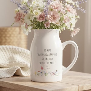 May include: A white ceramic jug filled with a bouquet of pink, white, and purple flowers. The jug features the text: To Mum, Wishing you a fabulous 60th birthday, Lots of love Katie x, with a small butterfly and floral graphic at the base.