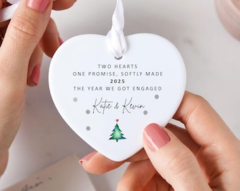May include: A white heart-shaped ornament with a white ribbon. The ornament has the text "TWO HEARTS ONE PROMISE, SOFTLY MADE 2025 THE YEAR WE GOT ENGAGED" and the names "Kate & Kevin". A small green Christmas tree is at the bottom.