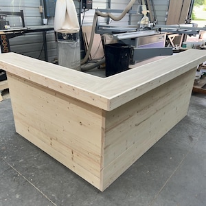 May include: A large, unfinished wooden bar counter with a light wood finish. The counter has a rectangular shape with a corner cut out, creating a L-shape. The counter is made of multiple planks of wood, creating a rustic look.