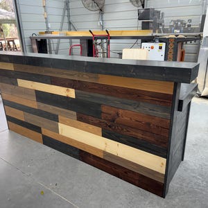 May include: A rustic wooden bar counter with a black countertop and a reclaimed wood front. The wood is a mix of light and dark brown, gray, and black.