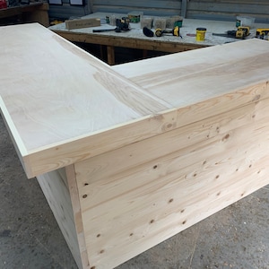 May include: A wooden L-shaped bar counter with a light wood top and a natural wood base. The top is made of plywood and has a light wood trim around the edge. The base is made of planks of wood and has a natural wood finish.