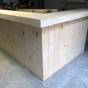 May include: A large, unfinished wooden bar with a rectangular countertop. The bar is made of light-colored wood planks and has a simple, rustic design.