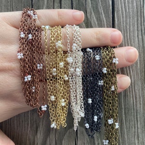 May include: A close-up of six different chain necklaces with small white beads. The chains are in copper, gold, silver, black, and two shades of brass.