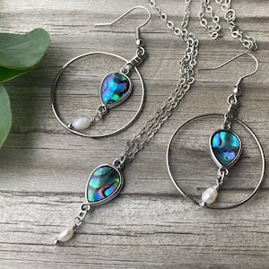 May include: A set of silver hoop earrings with a teardrop-shaped abalone shell pendant and a pearl accent. The earrings are paired with a matching necklace.