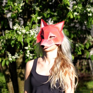 May include: A person wearing a red fox mask with a geometric design. The mask is made of paper and has a white background. The person is standing in front of a green bush with white flowers.