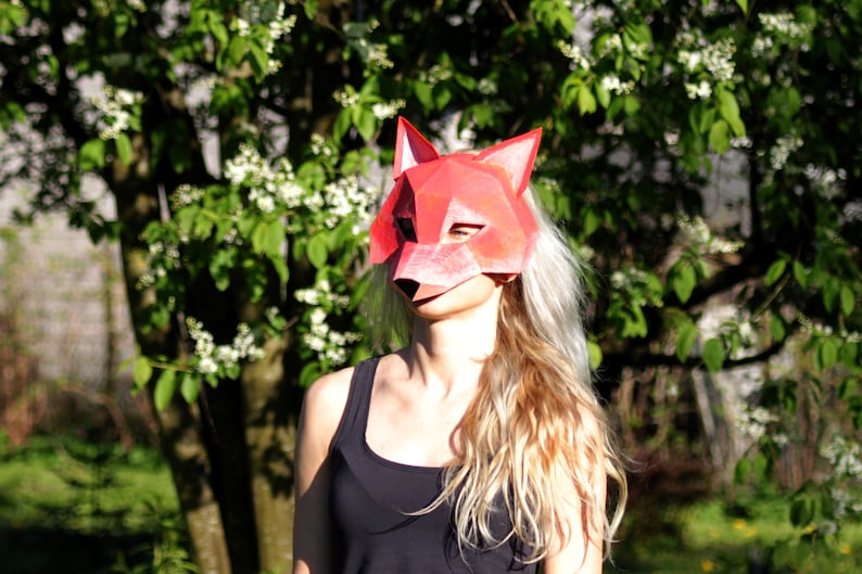 May include: A person wearing a red fox mask with a geometric design. The mask is made of paper and has a white background. The person is standing in front of a green bush with white flowers.
