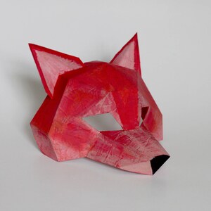 May include: A red paper fox mask with a geometric design. The mask has a black nose and eye holes.