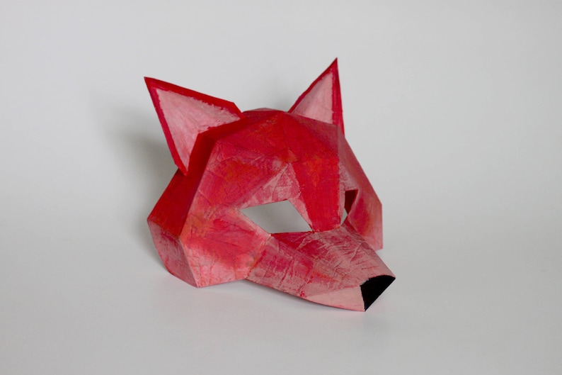 May include: A red paper fox mask with a geometric design. The mask has a black nose and eye holes.