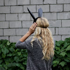 Puede incluir: Una mujer con una m&aacute;scara de conejo de papel negro con orejas grandes. Est&aacute; de pie frente a una pared de ladrillo blanco con plantas verdes en primer plano.