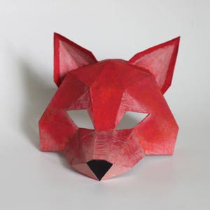 May include: A red paper mask shaped like a fox head. The mask has a black nose and eye holes.