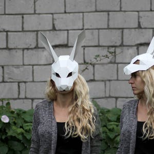Puede incluir: Dos personas con m&aacute;scaras de conejo de papel mach&eacute; blanco. Las m&aacute;scaras tienen orejas grandes y son de forma geom&eacute;trica. Las personas llevan su&eacute;teres grises y tienen el pelo largo y rubio.