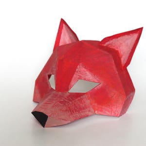 May include: A red paper fox mask with a geometric design. The mask has cut-out eye holes and a pointed nose.
