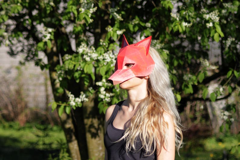 May include: A woman wearing a red fox mask made of paper. The mask has a pointed snout and large ears. The woman has long blonde hair and is wearing a black tank top.