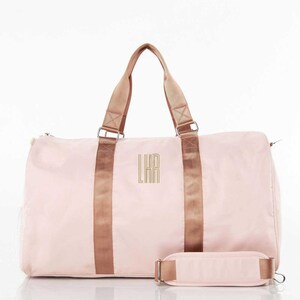 May include: A light pink duffel bag with brown straps and handles. The bag has a shoulder strap and a zipper closure. The initials "LKR" are embroidered in gold on the front of the bag. The bag is designed for travel or gym use.