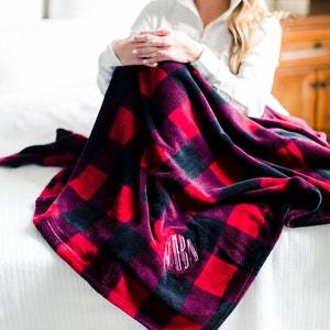 May include: A red and black plaid fleece blanket with a monogrammed initial in the center. The blanket is folded and draped over a white bed.