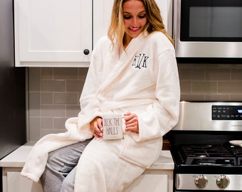 May include: A woman wearing a white robe with the initials "K.I.K." embroidered on the front, sits on a kitchen counter holding a white mug with the text "Deck the Halls".