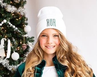 May include: A young person wearing a white beanie with the word "AUR" embroidered in green. They are wearing a green velvet jacket and have long blonde hair.