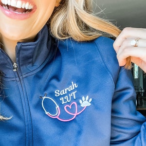 May include: A navy blue zip-up jacket with a white embroidered stethoscope, a pink heart, and a paw print. The text "Sarah LVT" is embroidered on the jacket.