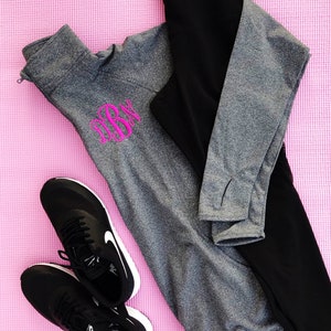 May include: A gray and black workout outfit with a pink monogram on the gray shirt. The outfit includes a long-sleeved shirt, leggings, and black running shoes.