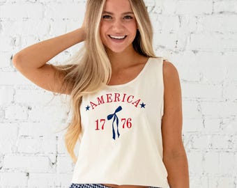 May include: Cream-colored tank top with the word "AMERICA" in red and blue, along with the year "1776". The tank top is paired with blue and white checkered boxer shorts. The outfit is casual and patriotic.