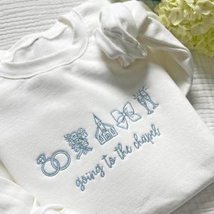 May include: White sweatshirt with light blue embroidered wedding-themed graphics, including rings, flowers, a church, a bow, and champagne glasses. The phrase "going to the chapel" is embroidered below the graphics.