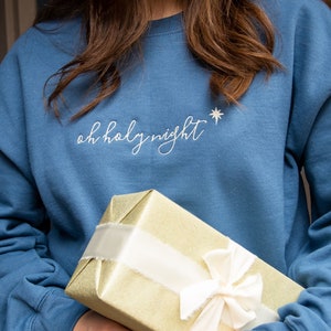 May include: A person wearing a blue sweatshirt with the text "oh holy night" embroidered on the front. The person is holding a gold gift box wrapped with a white ribbon.