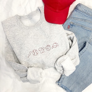 May include: A grey sweatshirt with a red embroidered baseball themed design. The design includes a baseball bat, baseball, jersey, glove, and baseball cap.