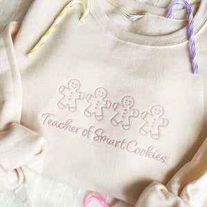 sweet cream crewneck sweatshirt with custom gingerbread men icons and "teacher of smart cookies" design embroidered in powder pink thread