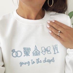 May include: White sweatshirt with light blue embroidered wedding-themed graphics, including rings, flowers, a church, a bow, and champagne glasses. The phrase "going to the chapel" is embroidered below the graphics.