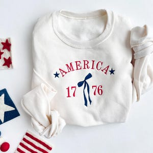 May include: Cream-colored sweatshirt with the word "AMERICA" arched across the chest in red, with blue stars on either side. Below, the number "1776" is embroidered in red. A blue bow and streamers are centered below the word "AMERICA".