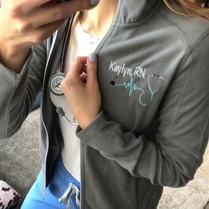 May include: Gray fleece jacket with a zipper and a stethoscope. The jacket has an embroidered design that says "Kaylyn, RN" with a heart beat and a stethoscope.
