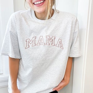 K&ouml;nnte beinhalten: Ein graues Oversize-T-Shirt mit dem Wort "MAMA" in rosa gestickten Buchstaben.