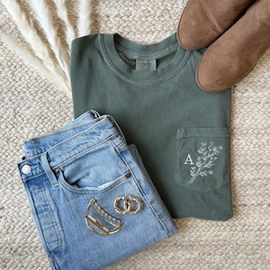 May include: An olive green pocket t-shirt with a white floral design and the letter "A" embroidered on the pocket. Light blue jeans, brown ankle boots, and gold earrings are also in the image. The items are arranged on a woven rug.