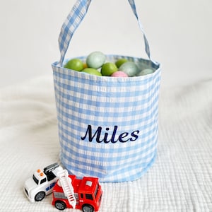 Peut inclure: Un panier de Pâques en gingham bleu et blanc avec un nom brodé en bleu "Miles" sur le devant. Le panier est rempli d'œufs de Pâques en plastique colorés. Deux camions jouets sont au premier plan.