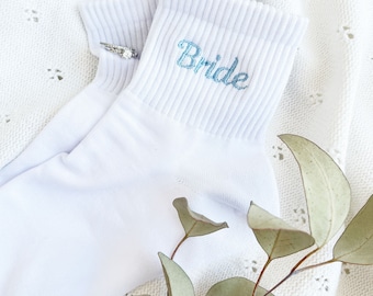 May include: White ankle socks with the word "Bride" embroidered in light blue on one sock. A silver ring is on the sock.