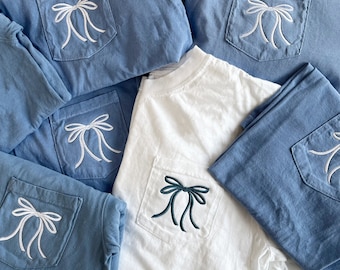 May include: Five t-shirts with embroidered bows on the pockets. Four of the shirts are blue denim with white bows. One shirt is white with a dark green bow.