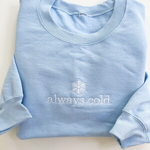 May include: A light blue sweatshirt with a white snowflake and the text "always cold" embroidered on the front.