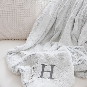 Pu&ograve; includere: Una morbida coperta in finta pelliccia grigio chiaro con una grande lettera "H" ricamata in grigio pi&ugrave; scuro. La coperta &egrave; piegata e drappeggiata su una superficie bianca, con un cuscino bianco strutturato sullo sfondo.